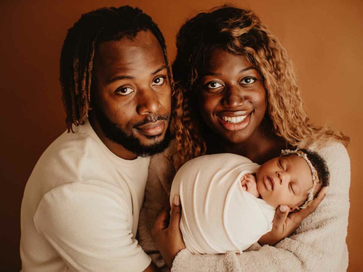 Casal posando para uma foto com seu recém-nascido. A mãe segura o bebê delicadamente, embrulhado em um paninho, enquanto o pai abraça a mãe com carinho.