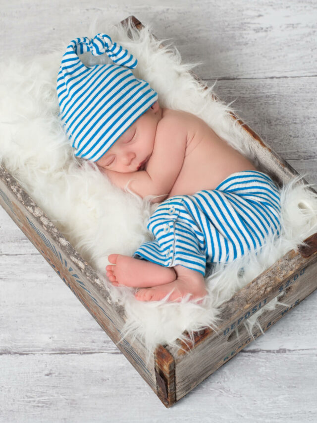 Bebê fazendo ensaio newborn, ele esta deitado dentro de uma caixa forrada com um pano peludinho. O neném usa uma calça e um gorro azul de listras.