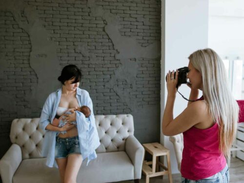 Fotógrafa realizando um ensaio newborn com todos os cuidados essenciais, garantindo conforto e segurança ao bebê. Ela segura uma câmera enquanto fotografa uma mãe amamentando seu recém-nascido.