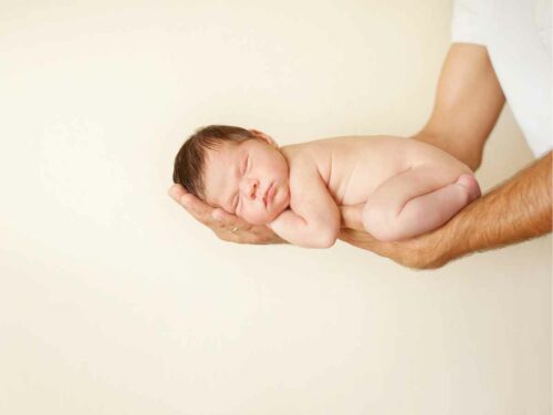 Pai segurando seu recém-nascido com delicadeza em uma posição que remete à postura fetal dentro do útero materno, durante um ensaio fotográfico estilo newborn.