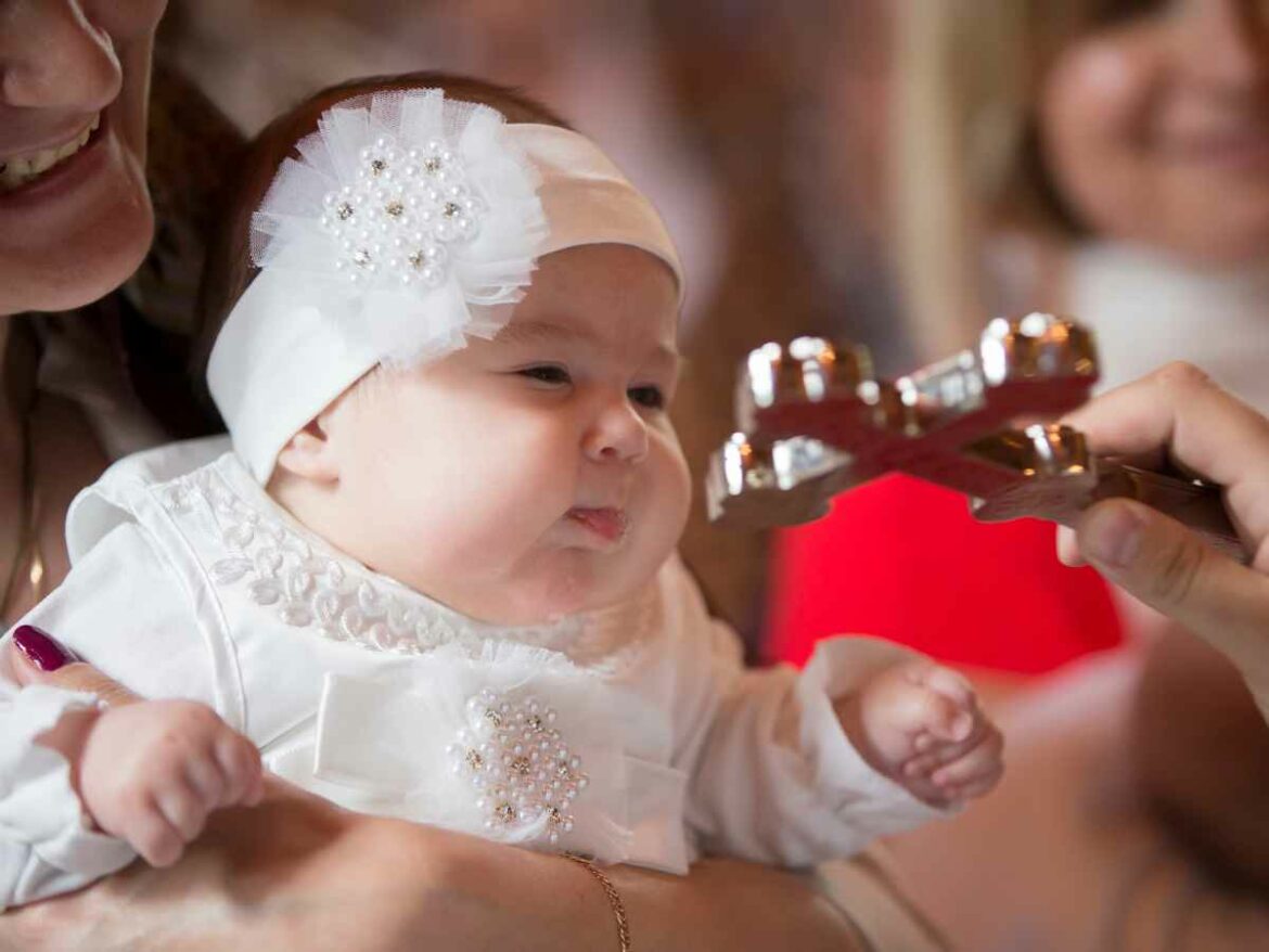 Bebê sendo batizado em uma igraja. A mãe segura sua filha que está vestida com roupas tipicas, enquanto o padre a abençoa com a cruz sagrada.