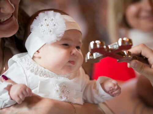 Bebê sendo batizado em uma igraja. A mãe segura sua filha que está vestida com roupas tipicas, enquanto o padre a abençoa com a cruz sagrada.