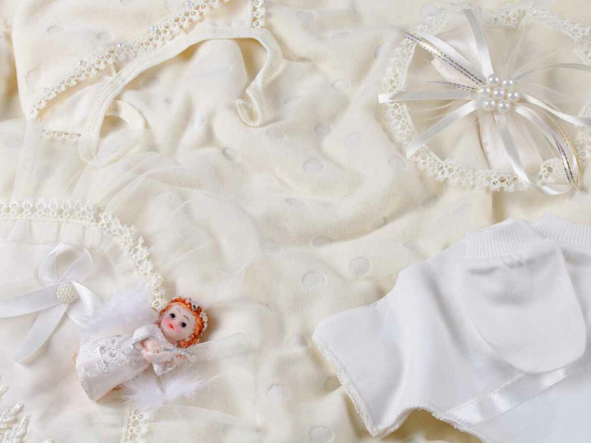 Roupas e acessórios delicados de batizado para bebê dispostos sobre um tecido branco com detalhes em renda. A composição inclui um vestidinho com tule, um par de meias, e um anjinho de enfeite.