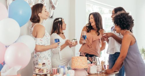 Mulheres celebrando em chá de bebês, sorrindo, segurando taças, com balões na decoração, bolo, cupcakes sobre a mesa... e simbolizando brincadeiras de chá de bebê