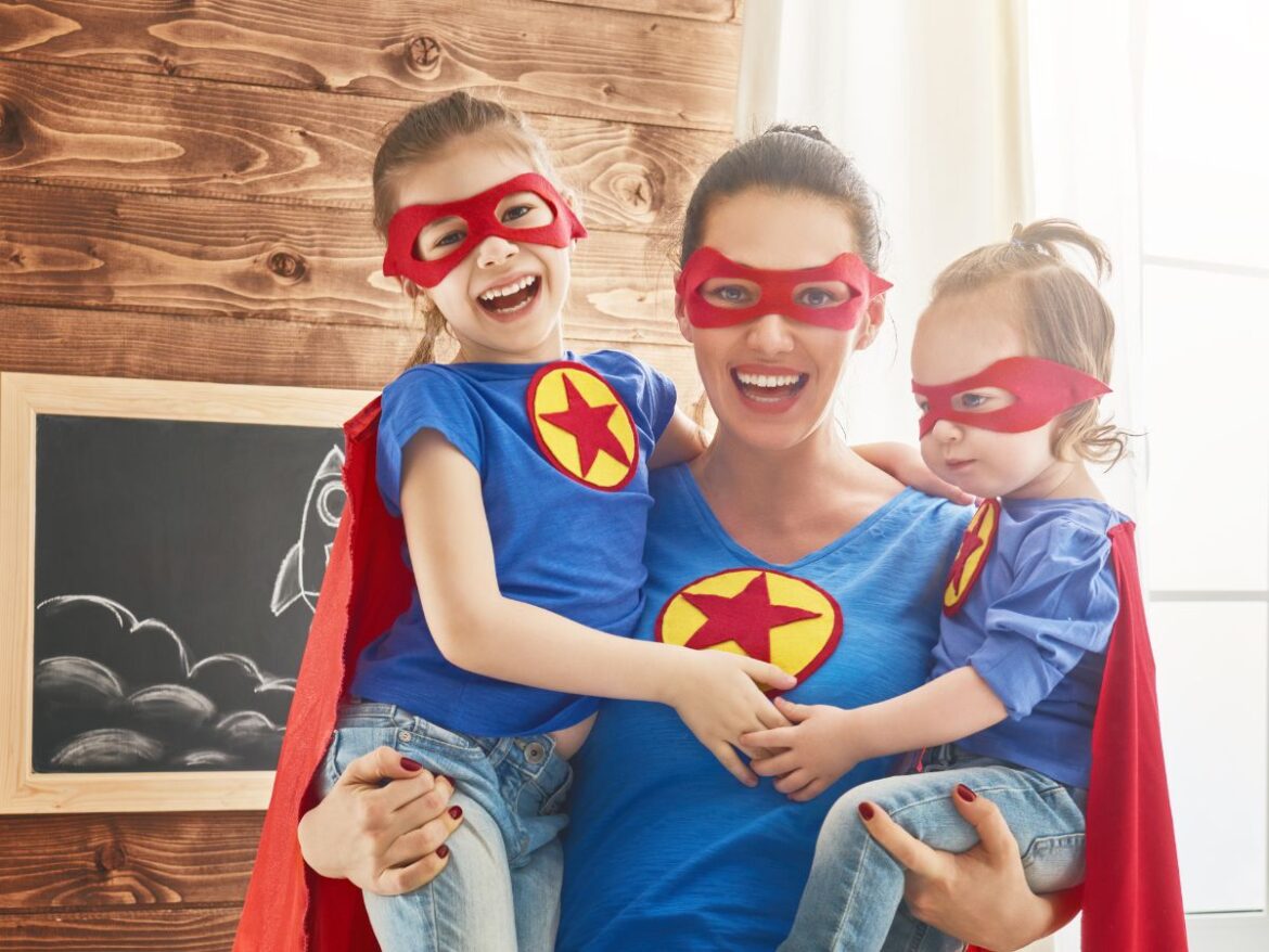 Mãe com duas filhas usando fantasias de super-heróis, com máscaras vermelhas, capas e camisetas azuis, em momento divertido em família.