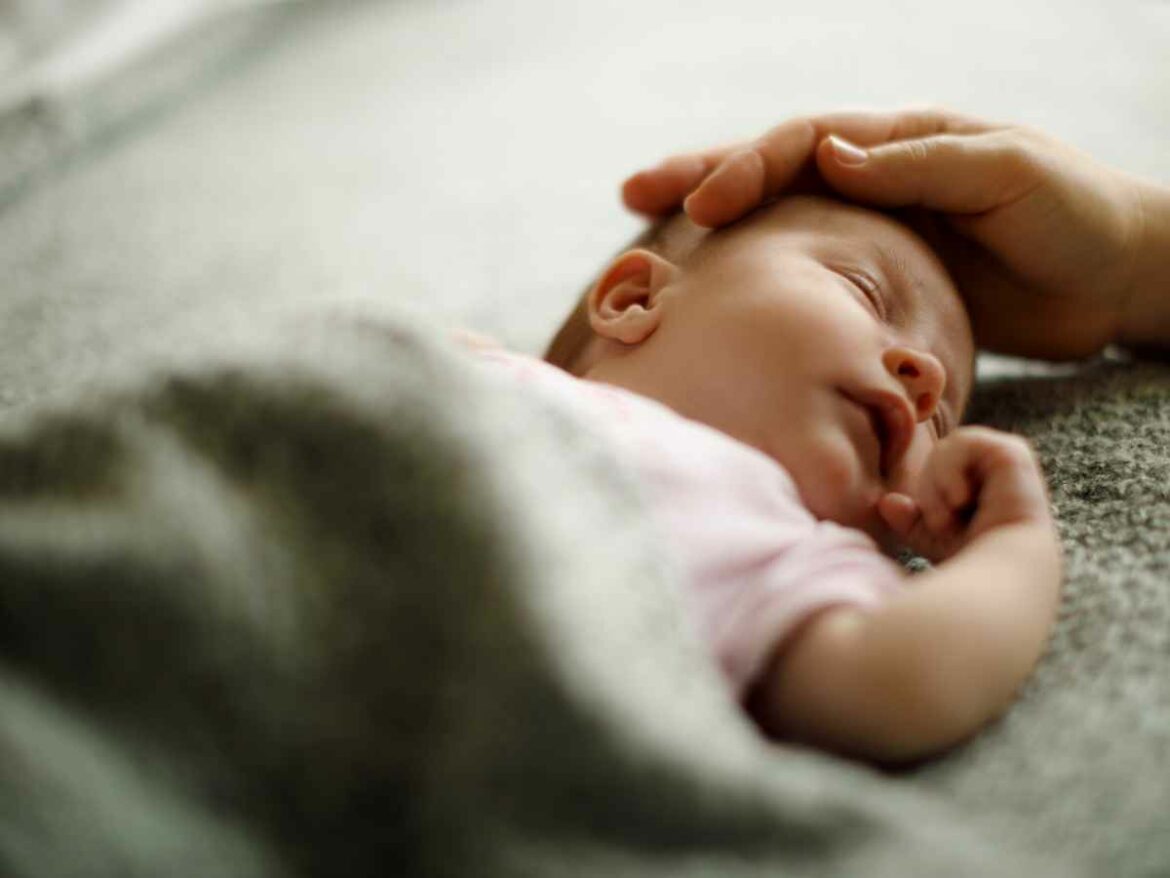 Bebê dorme tranquilamente deitado de lado sob um cobertor cinza, enquanto uma mão adulta acaricia suavemente sua cabeça.