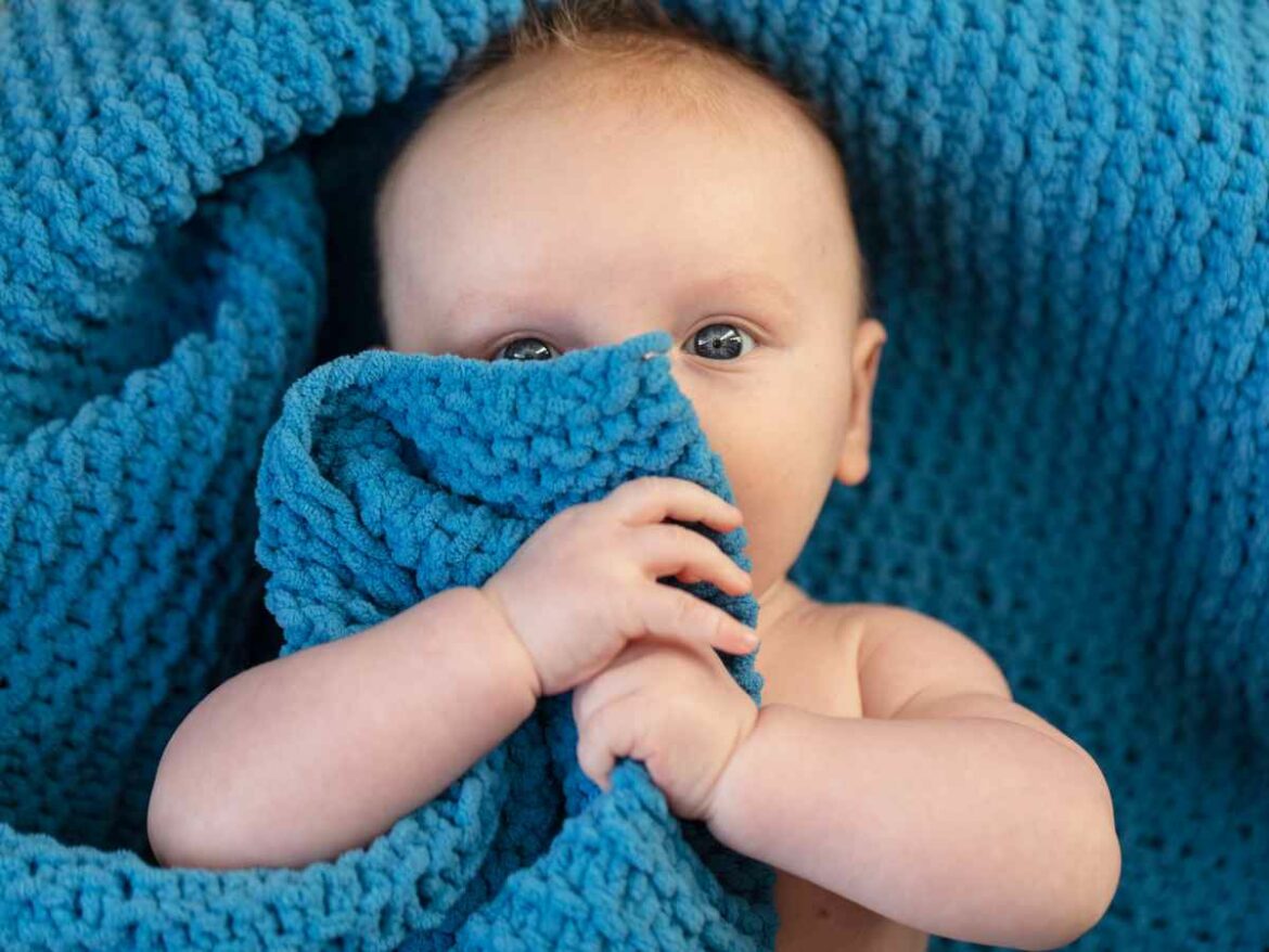Bebê de olhos claros parcialmente coberto por uma manta azul de crochê, segurando o tecido com as mãos enquanto observa atentamente.
