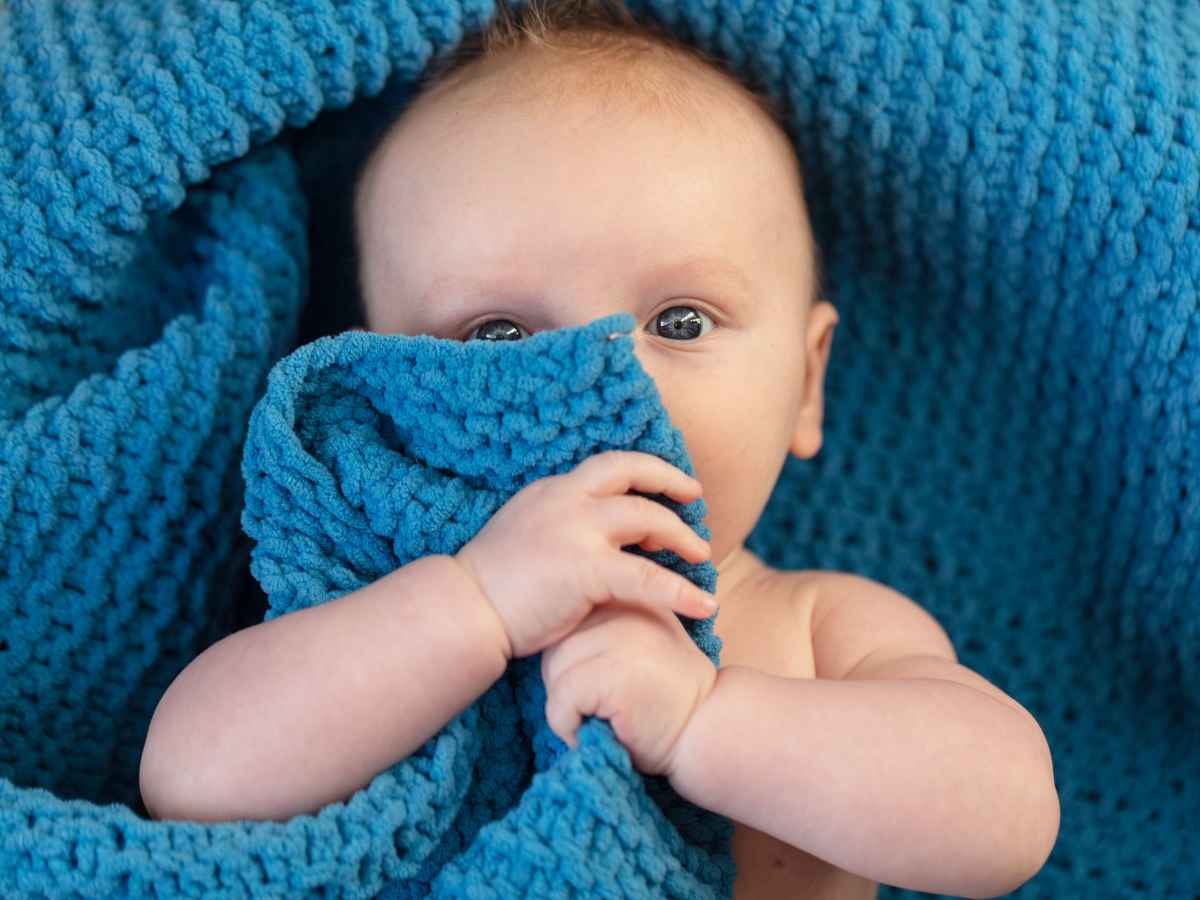 Bebê de olhos claros parcialmente coberto por uma manta azul de crochê, segurando o tecido com as mãos enquanto observa atentamente.