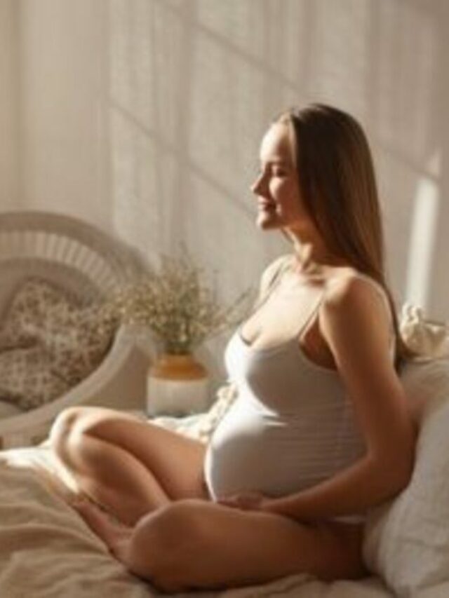 Foto de gestante em casa sentada sobre a cama de forma descontraída com luz natural. Emoções maternas.