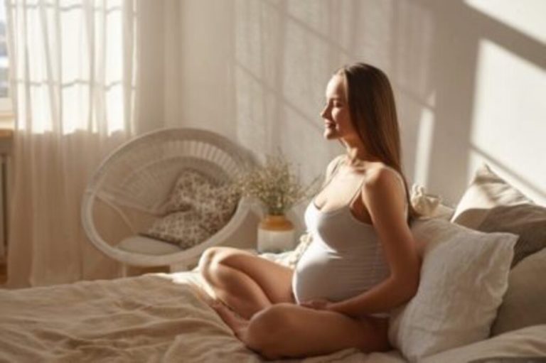 Foto de gestante em casa sentada sobre a cama de forma descontraída com luz natural. Emoções maternas.