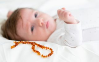 Bebê de olhos azuis e pele clara e roupinha branca ao deitado ao lado de um colar de âmbar