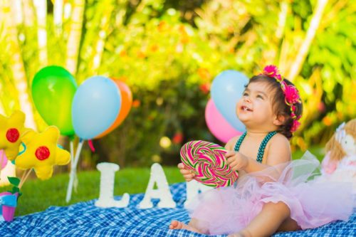 Bebê menina com tiara e segurando pirulito colorido, olhando para o céu rindo, com saia fuê em ambiente externo de jardim e balões e flores de pelúcia ao redor da bebê, simbolizando ideia para fotos de mesversário menina