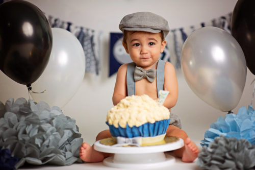 Bebezinho com bolo em sua frente, gravatinha e boina, com balões ao redor, simbolizando ideia para fotos de mesversario