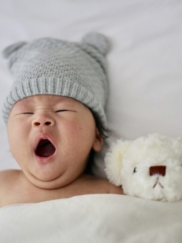 Bebê bocejando de touquinha ao lado de urso de pelúcia no berço indicando a janela de sono