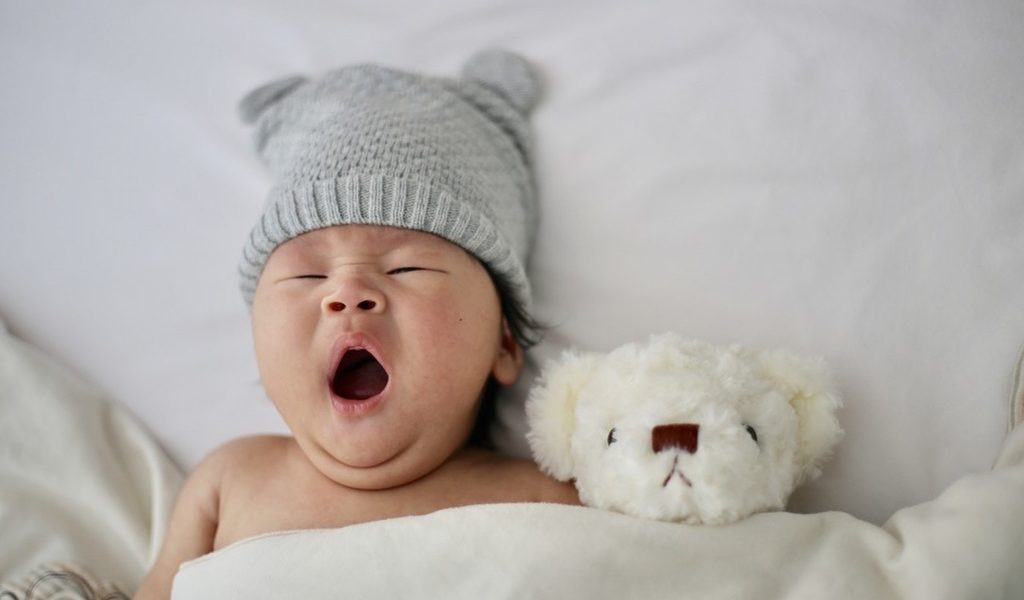Bebê bocejando de touquinha ao lado de urso de pelúcia no berço indicando a janela de sono