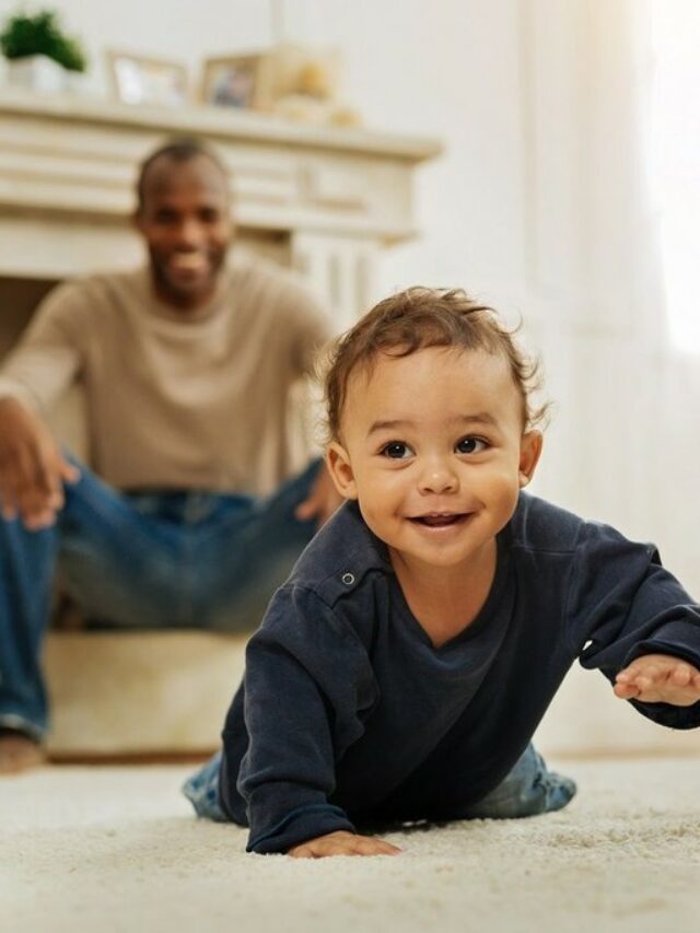 Pai ao fundo sorrindo agachado ajudando e estimulando o filho bebê, e bebê em primeiro plano engatinhando sorrindo, representando com qual idade o bebê começa a engatinhar