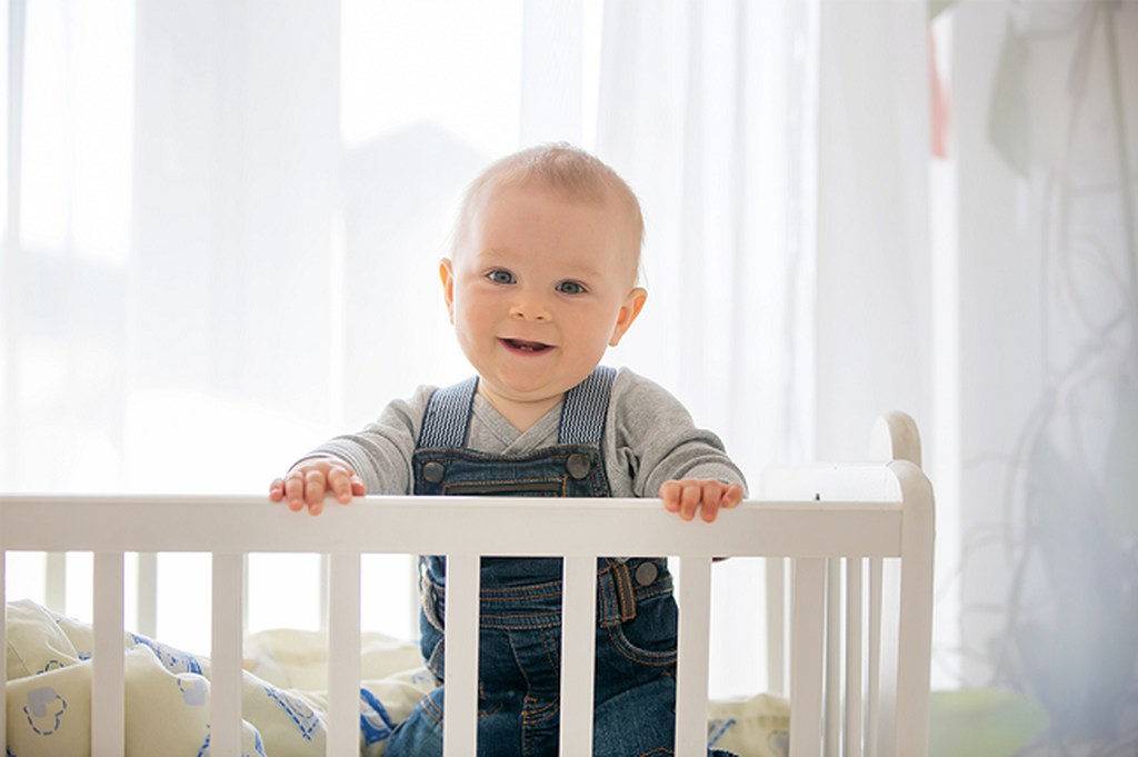 Bebê em pé, segurando na grade de segurança do berço, representando dermatite atópica em bebe