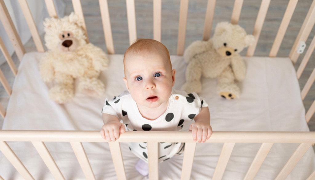 Bebê em pé no berço, olhando para cima com carinha de choro, simbolizando dermatite em bebe. Dois ursinhos de pelúcia ao fundo, dentro do berço