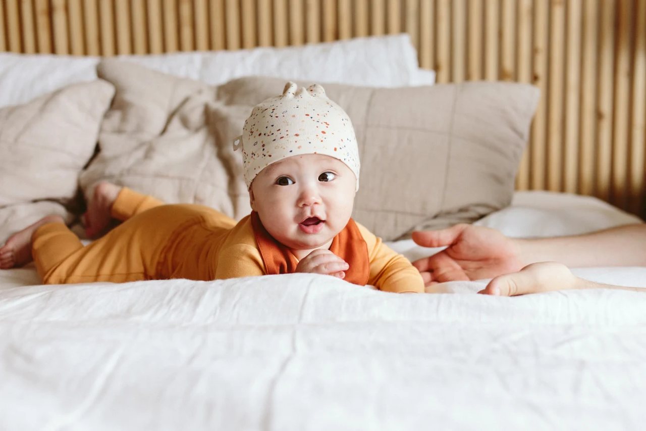 Bebê deitado de bruços na cama, olhando para a câmera, está usando uma touquinha e um macacão laranja