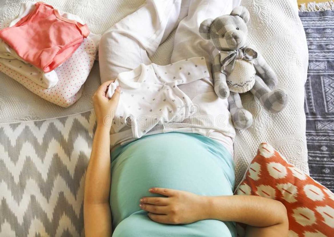 Grávida deitada, com roupinhas de bebê em seu colo, simbolizando o que levar na bolsa do bebe