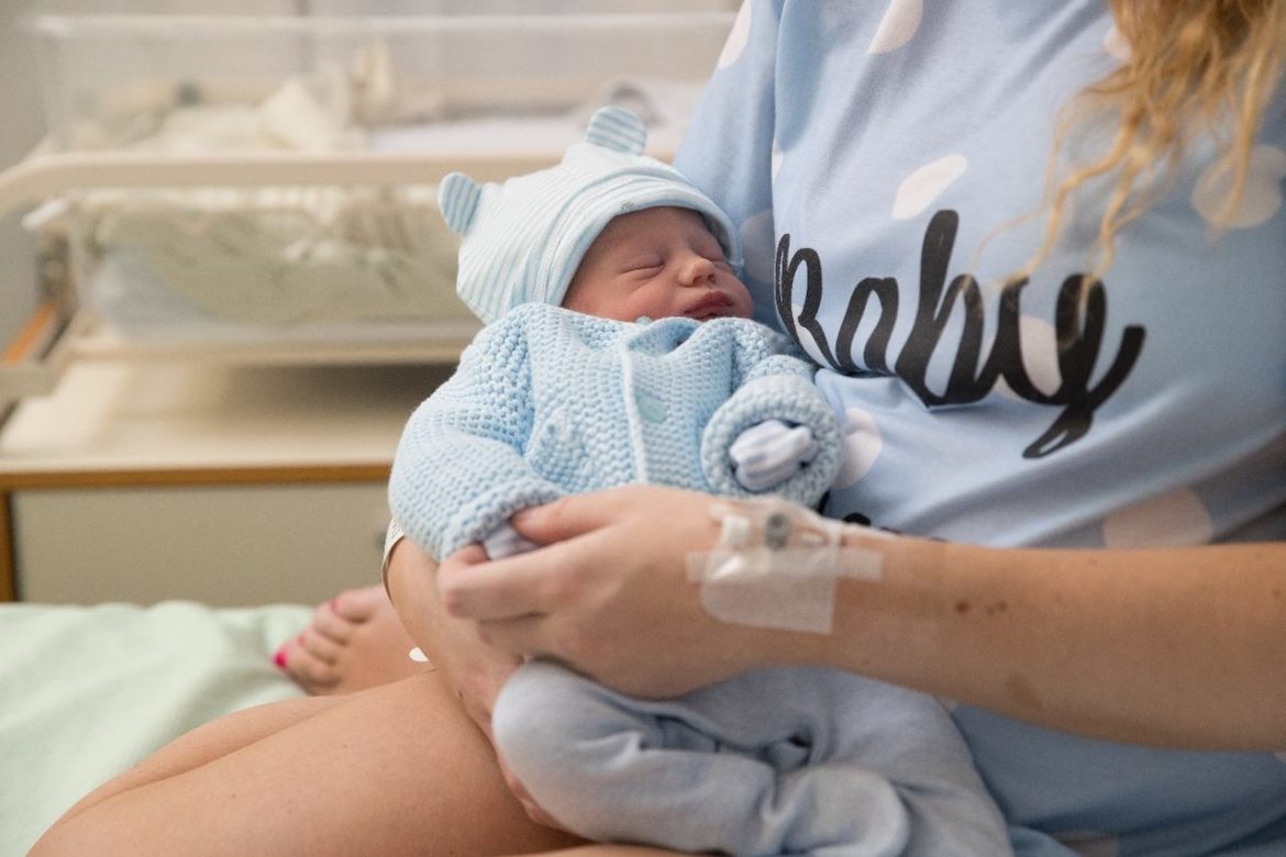 Mamãe com acesso de soro na mão segurando bebê recém-nascido na maternidade, o bebês está vestindo touquinha , luvas, calça e casaquinho de frio, todas as peças em azul, representando a primeira roupa do bebê ao nascer