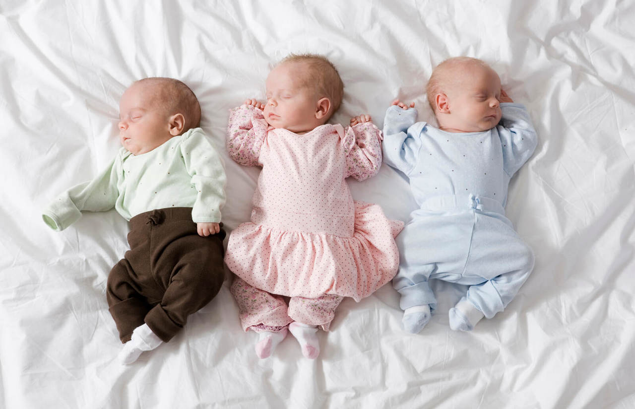Bebês trigêmeos deitados na cama, um deles com calça marrom e blusa de manga comprida verde clarinho, uma bebê de vestidinho rosa, e um bebê com conjuntinho azul claro. Os três bebês estão usando meias