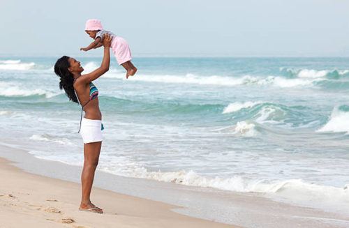 Mãe brincando com bebê na praia, mar ao fundo