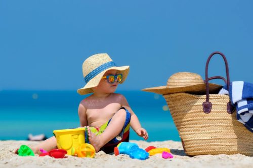 Bebê na praia de chapeuzinho e óculos escuros, com brinquedos com baldinho ao redor, olhando para sacola de praia da mãe, que contém chapéu e toalha, simbolizando cuidados com o bebe na praia e o que levar na bolsa