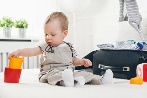 Bebê brincando com copos ou baldinhos coloridos ao lado de uma mala para viagem, na qual é possível ver um body cinza e sapatinhos