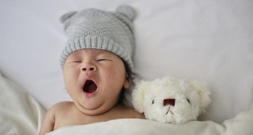 Bebê bocejando com touca de orelhinhas, deitado ao lado de ursinho de pelúcia, representando ideias de nomes para bebe 