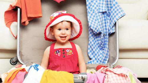 Bebê sorrindo com chapeuzinho dentro de uma mala para viagem aberta, com roupas espalhadas, simbolizando o que levar na mala de bebê para viagem