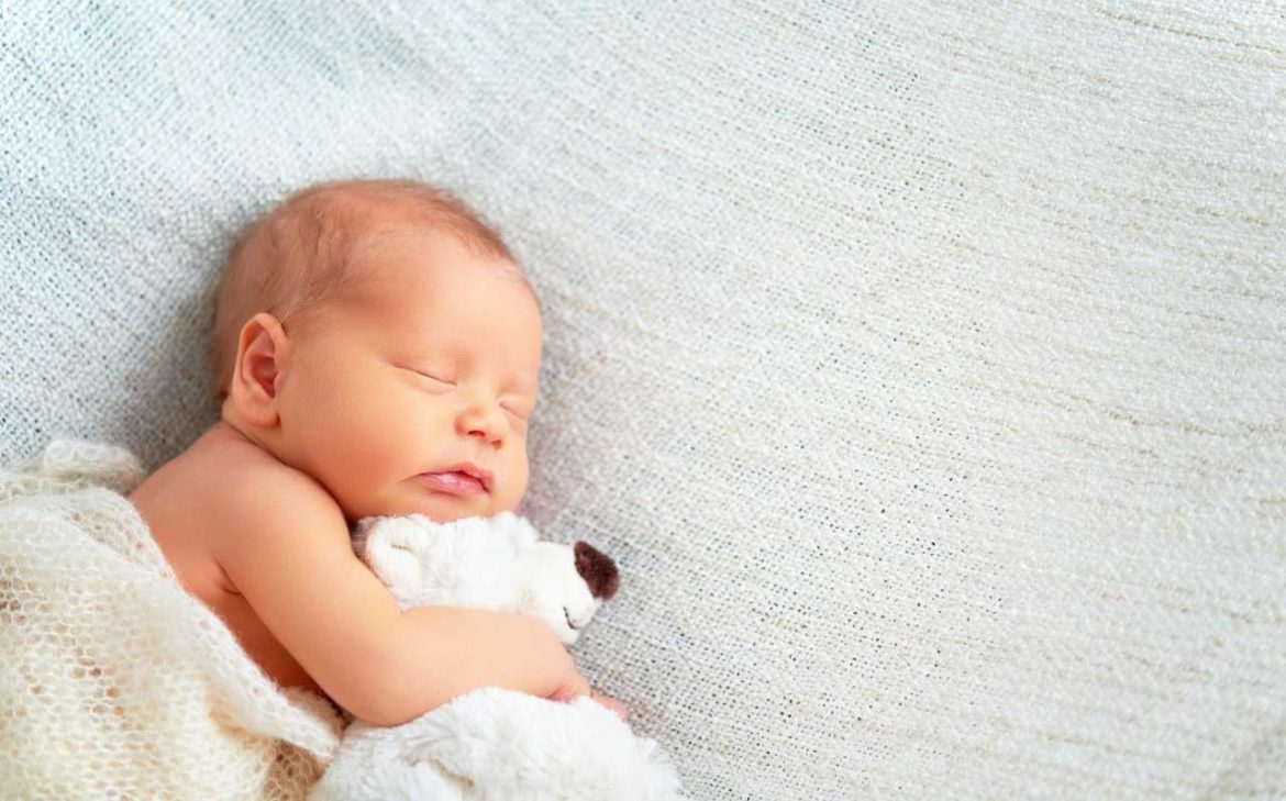 Bebê dormindo abraçando ursinho de pelúcia e coberto com manta, representando presente para bebe recém-nascido