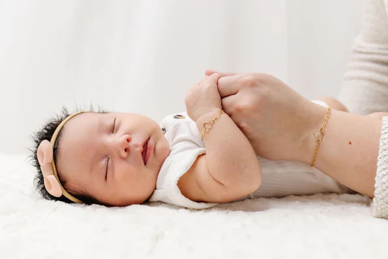 Mão segurando a mãozinha de um RN dormindo, e no pulso do bebê há uma joia para recém nascido