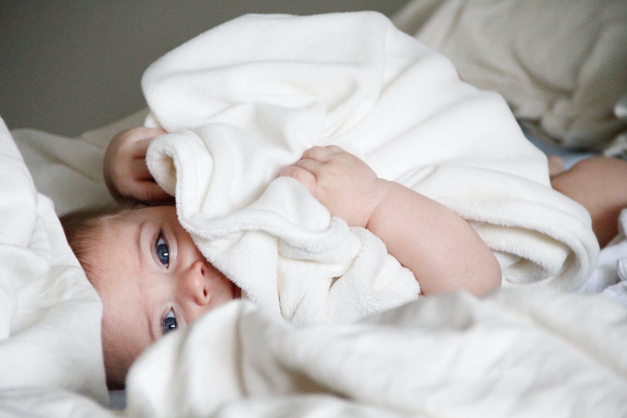Bebê deitado segurando manta branca ou toalha branca, simbolizando o que comprar para higiene do bebê recém nascido