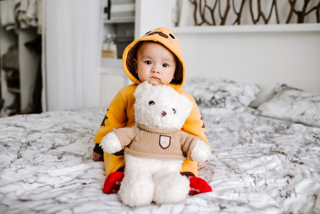 Bebê sentado na cama de touca e com ursinho de pelúcia, simbolizando cuidados com bebê no inverno
