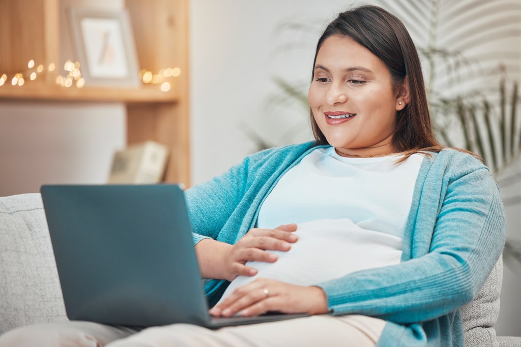 Gestante sorrindo sentada confortavelmente num sofá, com uma mão na barriga, e a outra usando notebook, simbolizando enxoval de bebe online