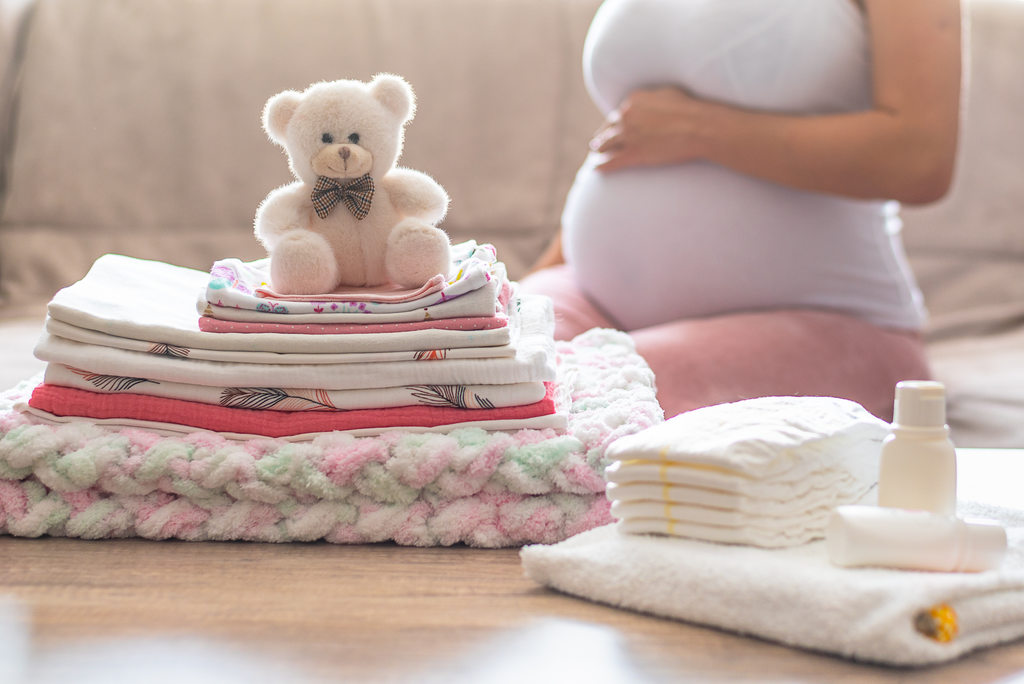 Simbolizando lista de roupas para recém nascido, gestante sentada segurando a barriga com toalha, lençóis, fronhas, manta e fraldas dobrados em sua frente. Além de um ursinho de pelúcia sobre a pilha de lençóis dobrados.