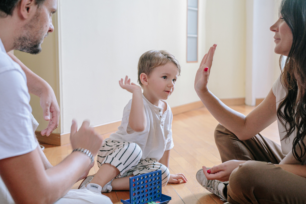 Mãe a esquerda, pai a direita e criança no centro, batendo as mãos No meio um brinquedo de colocar as peças