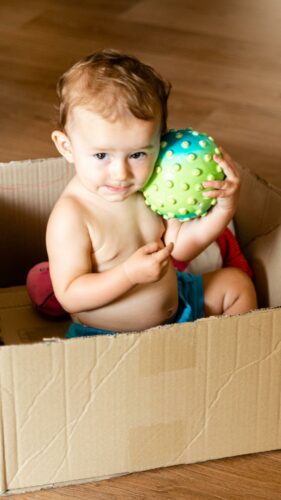 Bebê brincando dentro de uma caixa de papelão com uma bola em sua mão.