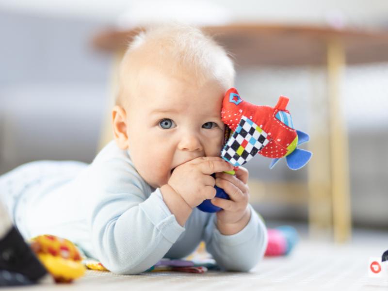 bebê deitado brincando com brinquedo colorido e olhando para a camera e outros brinquedos desfocados ao lado