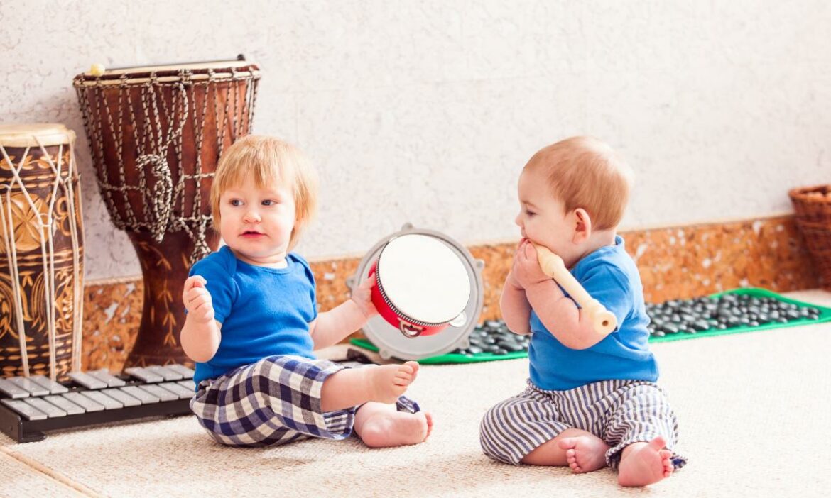 Dois bebês sentados no chão com instrumentos musicais em suas mãos, e outros instrumentos ao fundo