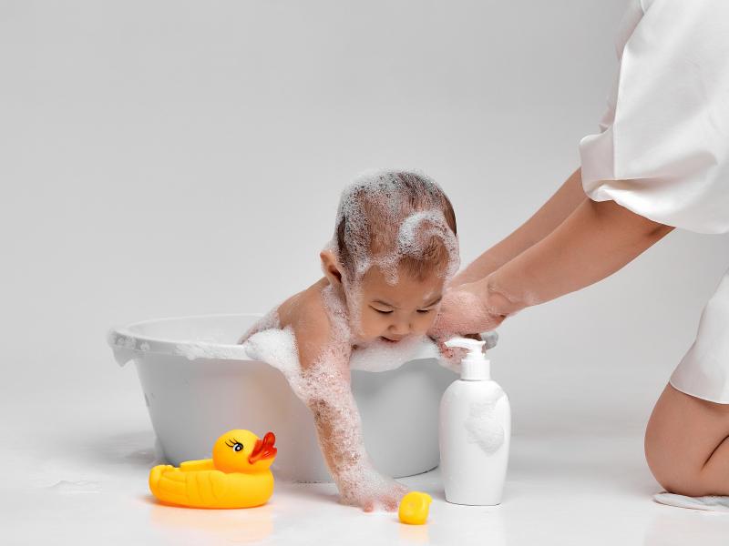 bebe tomando banho na banheira com o auxílio de um adulto e brinquedos e um frasco de shampoo ao lado da banheira