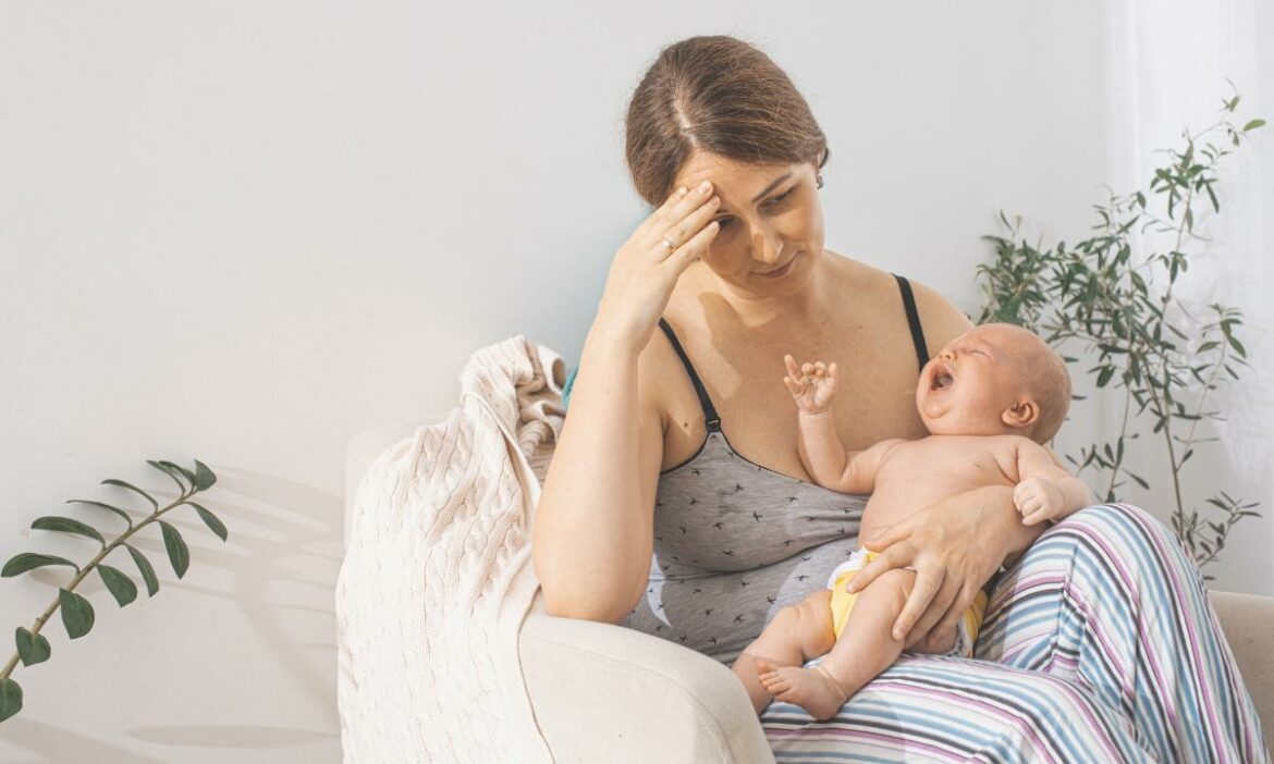 Mãe sentada, seu bebê em seu colo, e ela expressa dor ao realizar a amamentação