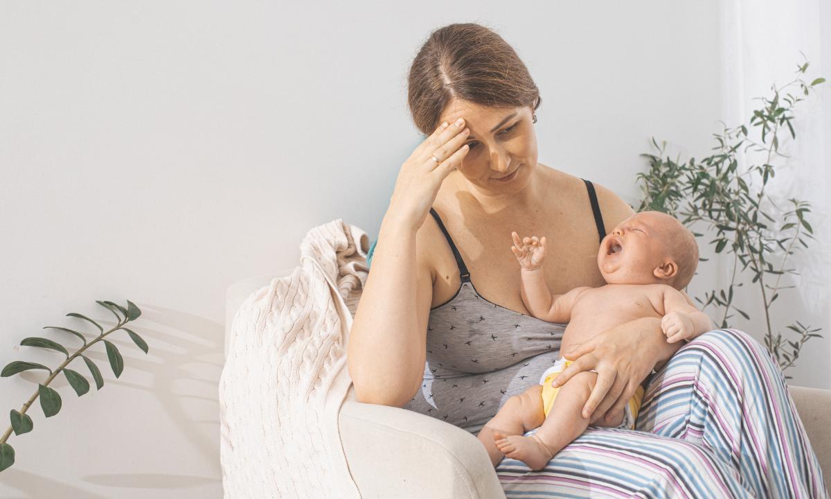 Mãe sentada, seu bebê em seu colo, e ela expressa dor ao amamentá-lo. Alimentos que podem causar cólicas no bebê.