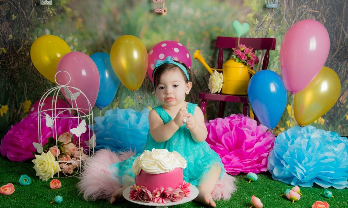 bebe sentada, a sua frente um bolo cor de rosa, e ao fundo, várias bexigas e outras decorações de festa de aniversário