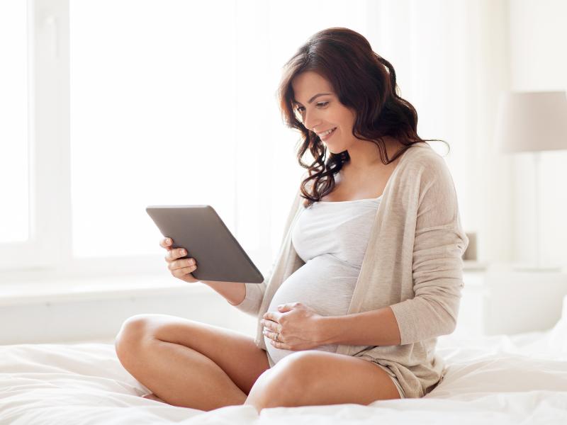 mulher grávida sentada com as pernas cruzadas olhando o tablet e sorrindo
