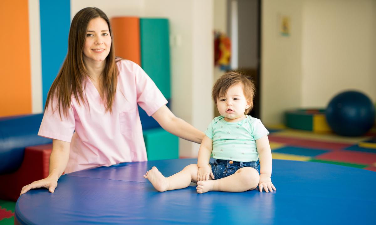 Bebê sentado em uma plataforma azul e uma mulher so lado estimulando seu desenvolvimento, formas coloridas ao fundo.