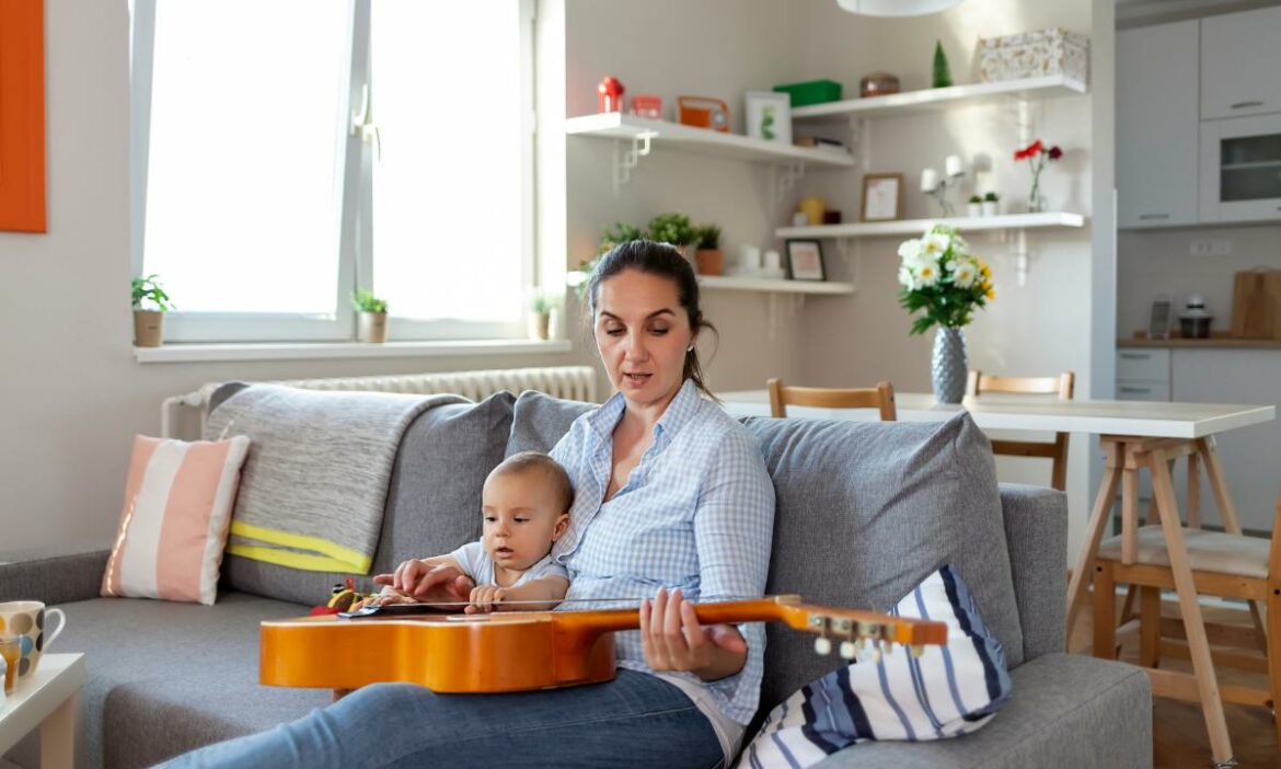 Mãe aplicando a música no desenvolvimento da sua criança, sentada no sofá com filho no colo tocando violão, mesa com flores ao fundo e outros objetos de decoração