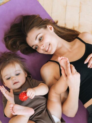 mãe e bebê deitadas e a bebê brinca com uma bola de massagear vermelha