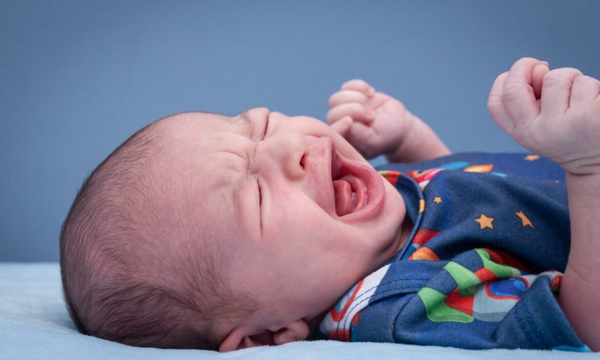 Bebê deitado com uma roupa azul com detalhes coloridos, chorando bastante